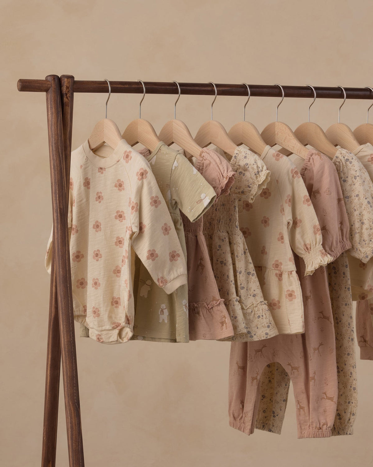 A wooden clothing rack holding multiple soft pastel baby outfits, including long-sleeve tops and pants, in cream, blush, and dusty pink tones with floral and animal prints, against a warm beige wall.