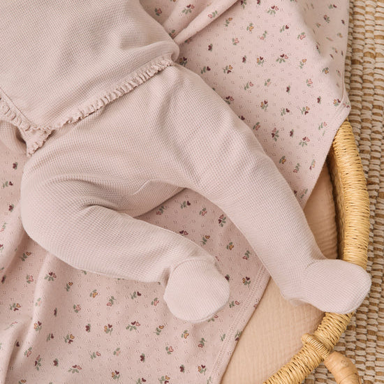 A close-up of a baby's legs wearing a pale pink waffle-knit footed sleeper, lying on a matching light pink floral blanket inside a woven rattan bassinet.