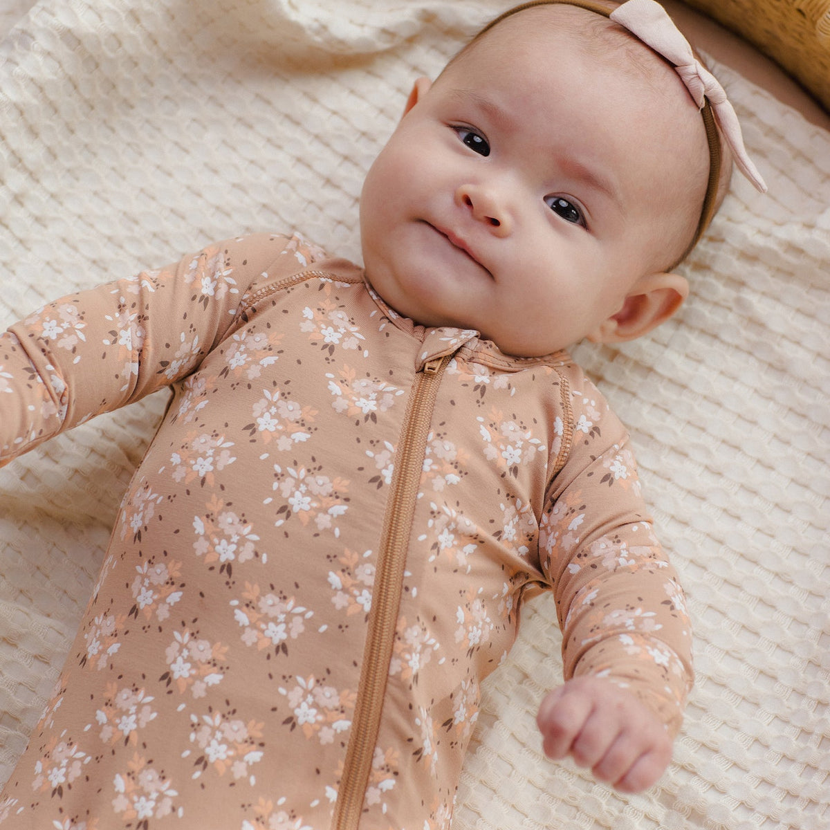 A baby lying on a textured cream blanket, wearing a long-sleeve beige floral zip-up sleeper and a matching pink bow headband.