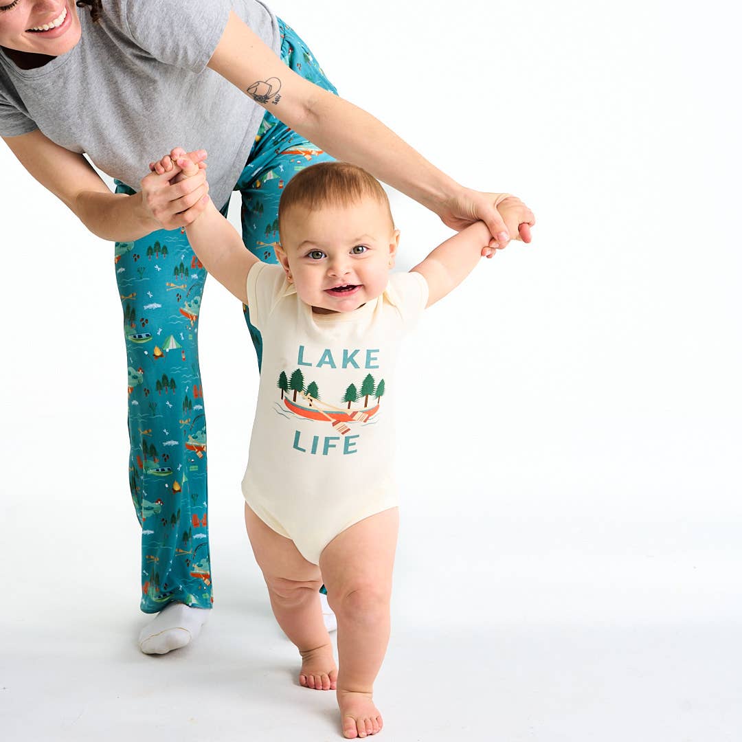 Baby wearing a 'Lake Life' onesie being helped to walk by an adult on a white background
