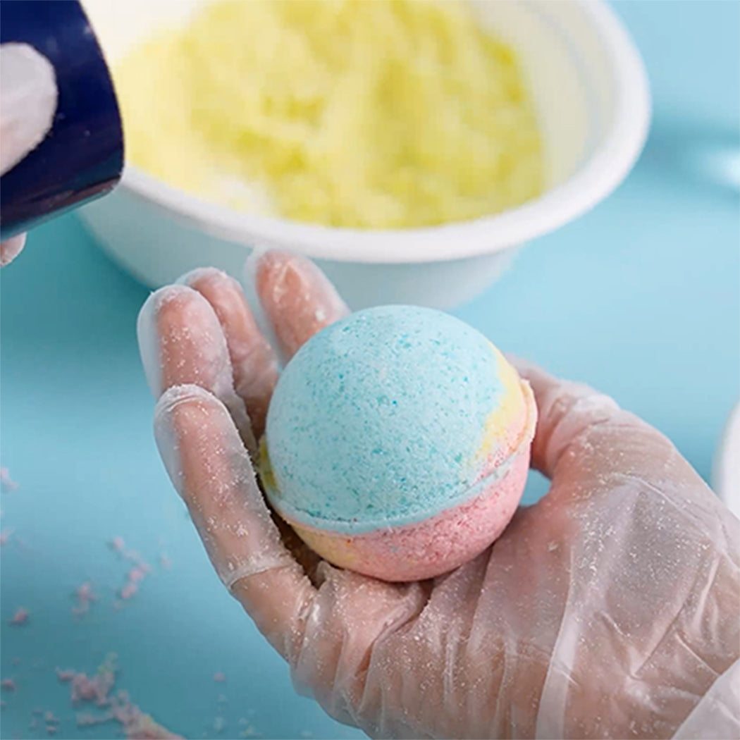 A hand wearing a clear plastic glove holds a colorful round bath bomb with a light blue top and pink bottom layers. A white bowl filled with yellow powder sits blurred in the background on a light blue surface.