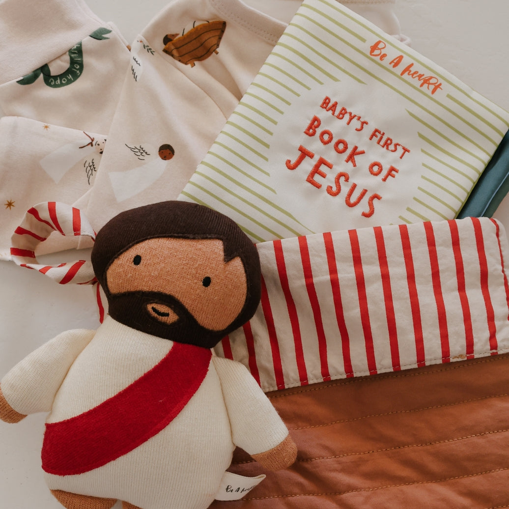 Plush toy of a child dressed as Jesus with a book titled 'Baby's First Book of Jesus' on a bed.