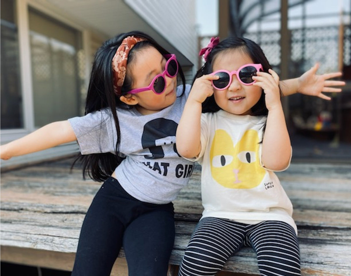 Two young girls sitting on a wooden bench wearing sunglasses and casual clothing.