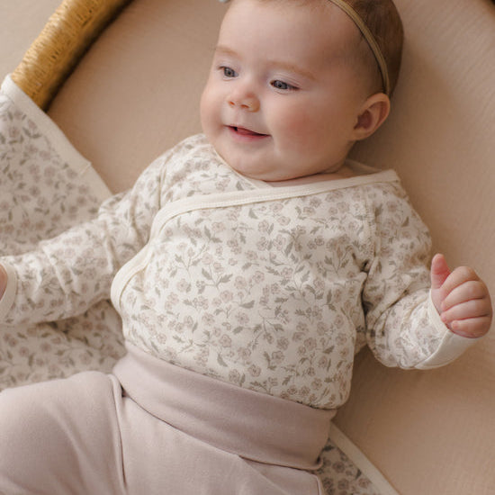 A baby lying on a woven bassinet wearing a cream floral long-sleeve wrap bodysuit with matching pants and a light gray bow headband.