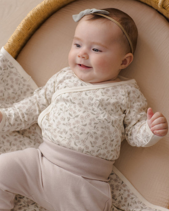 A baby lying on a woven bassinet wearing a cream floral long-sleeve wrap bodysuit with matching pants and a light gray bow headband.