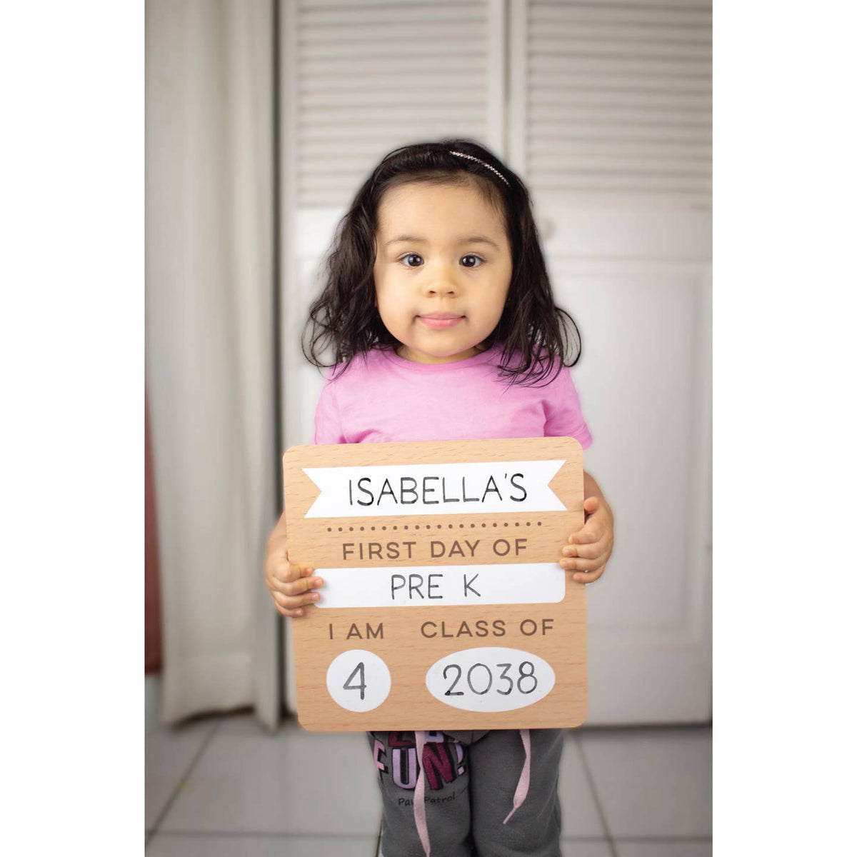 Child holding a sign for their first day of pre-K, with text on it.