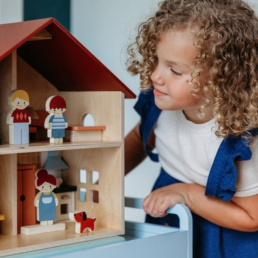 A young child with curly brown hair looking at an open-faced wooden dollhouse by Mentari, arranged on a white table with miniature wooden furniture and small figurines.