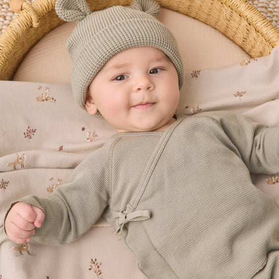 A baby lying on a woven mat inside a basket, wearing a sage green knit bonnet with two small pom-poms and a matching sage green long-sleeve romper. The baby is resting on a beige blanket with a small deer pattern.