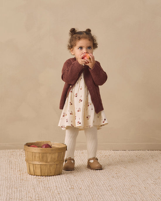 A toddler girl stands in a studio setting wearing a dark brown knit cardigan over a cream dress with a small floral print, white tights, and brown shoes. She is holding a red apple next to a small wooden basket on a textured cream rug against a warm beige backdrop.