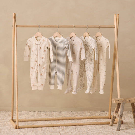 Five baby footed onesies in neutral tones hang on a wooden clothing rack against a beige wall and textured rug. The onesies include cream, gray-and-white stripes, and light floral and polka dot patterns.