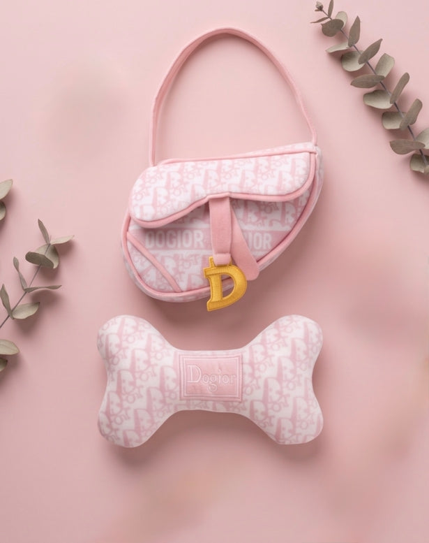 A pink Christian Dior dog harness bag and matching bone-shaped dog toy, both featuring the Dior Oblique monogram pattern, arranged on a pink background with decorative eucalyptus branches.