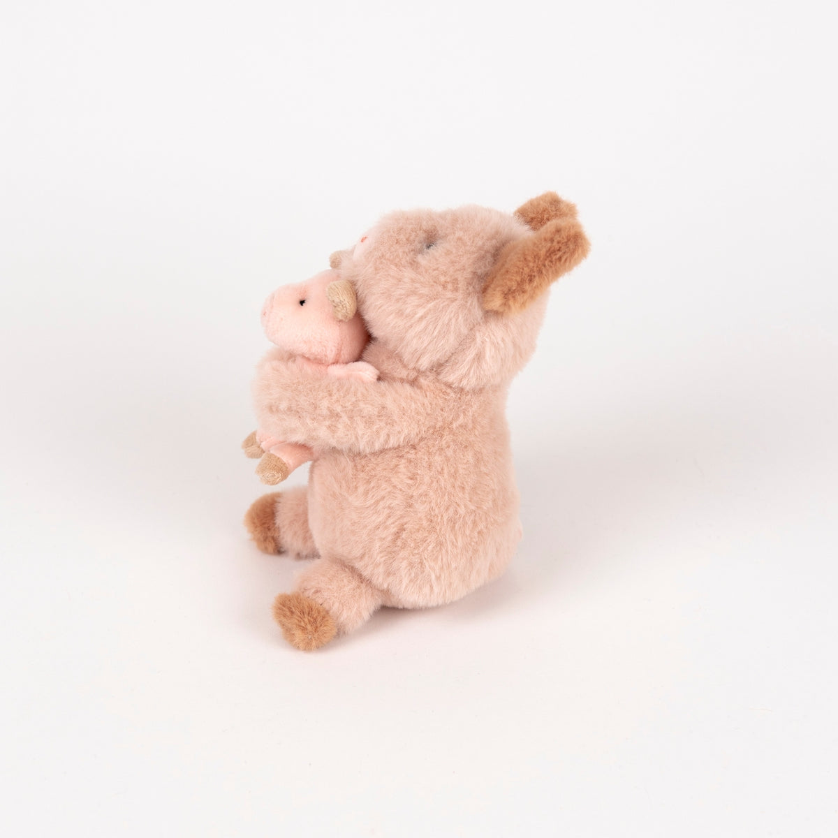 A plush stuffed animal featuring a light pink body and ears with darker brown accents, sitting and holding a small matching pink pig toy against a plain white background.