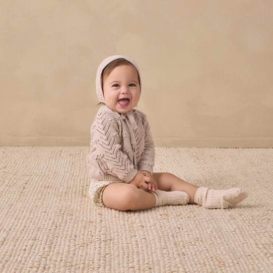 A baby sitting on a woven beige rug against a textured beige wall, wearing a cream cable-knit romper, matching bonnet, and knit socks.