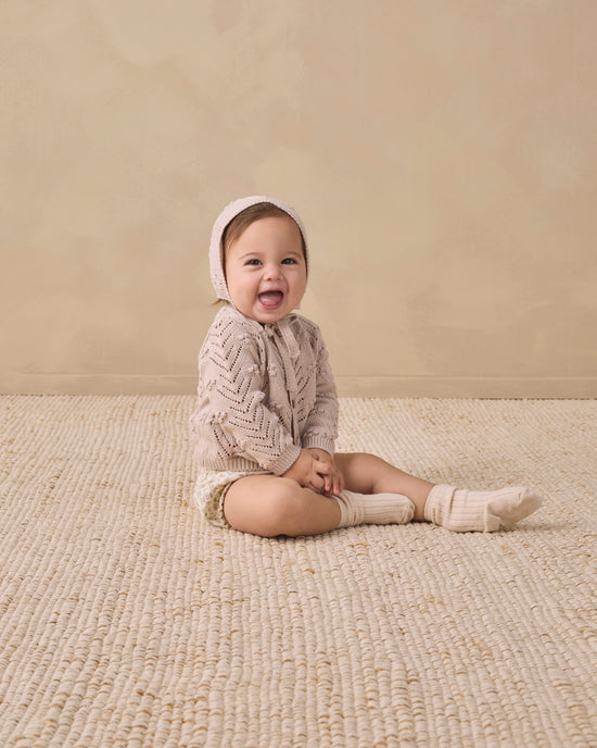 A baby sitting on a woven beige rug against a textured beige wall, wearing a cream cable-knit romper, matching bonnet, and knit socks.