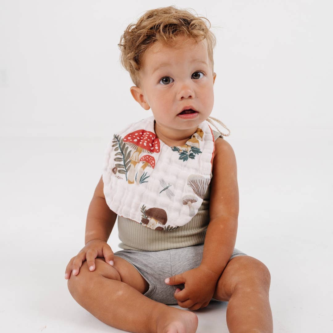 A toddler sitting on a white studio floor wearing a white bib with a colorful forest print of mushrooms, leaves, and a squirrel, paired with a gray ribbed tank top and shorts.