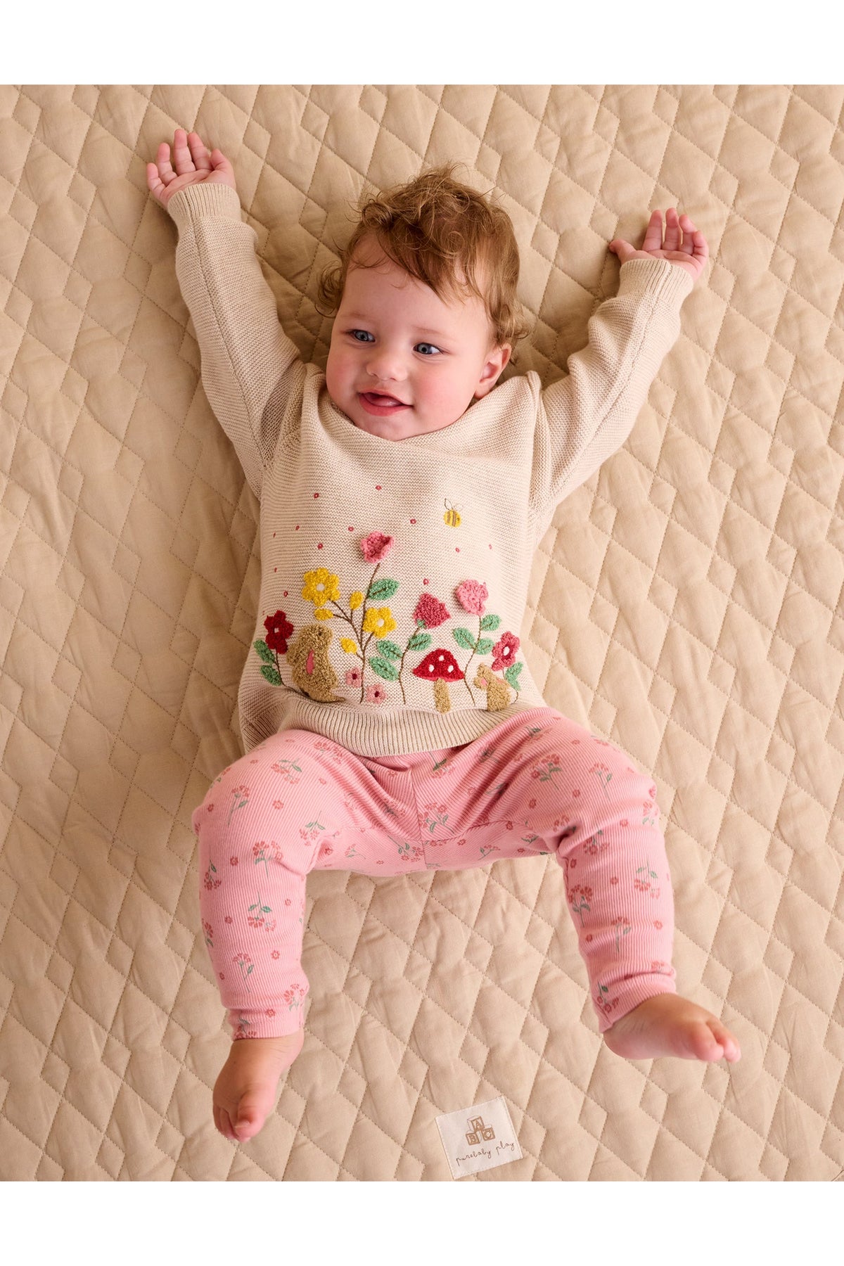 A baby lying on a quilted beige mattress, wearing a cream knit sweater with a colorful mushroom and flower design and pink patterned leggings.
