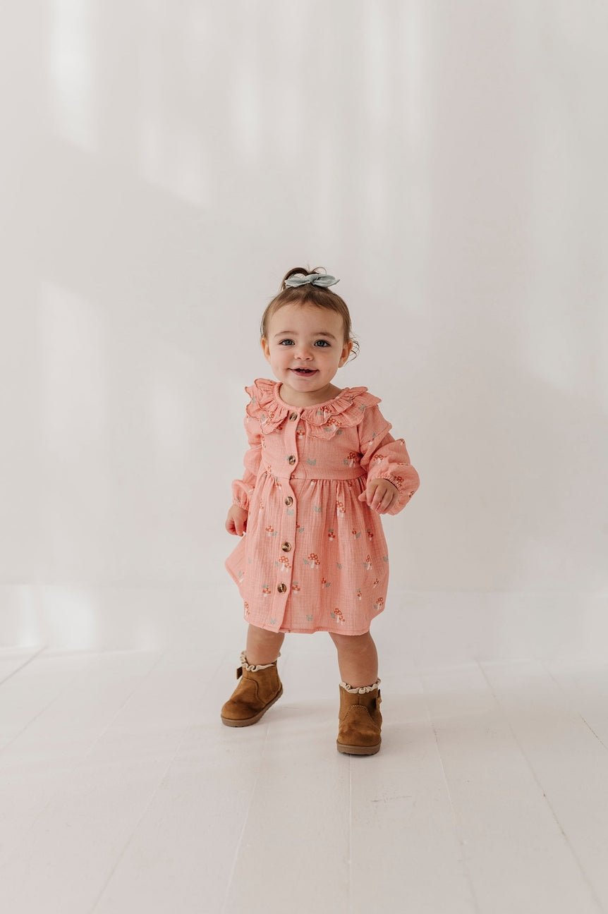 A toddler girl wearing a dusty pink long-sleeve button-up dress with a ruffled collar and small floral print, paired with brown boots, standing against a bright white studio background.