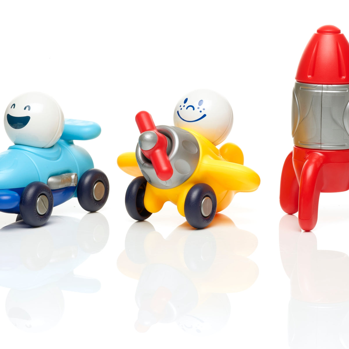 Three colorful plastic toy vehicles with smiling cartoon faces: a blue race car, a yellow airplane, and a red rocket ship, arranged on a white reflective surface against a white background.