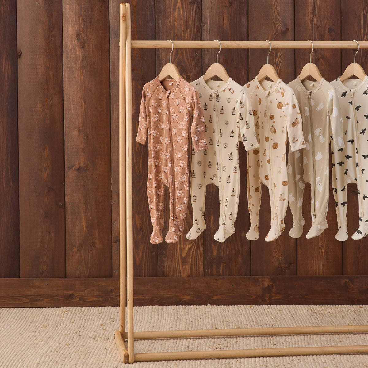 Five baby footed onesies in soft neutral tones hang on a wooden clothing rack against a dark wood plank wall. The onesies include pink floral, cream tree prints, and cream polka dot patterns.
