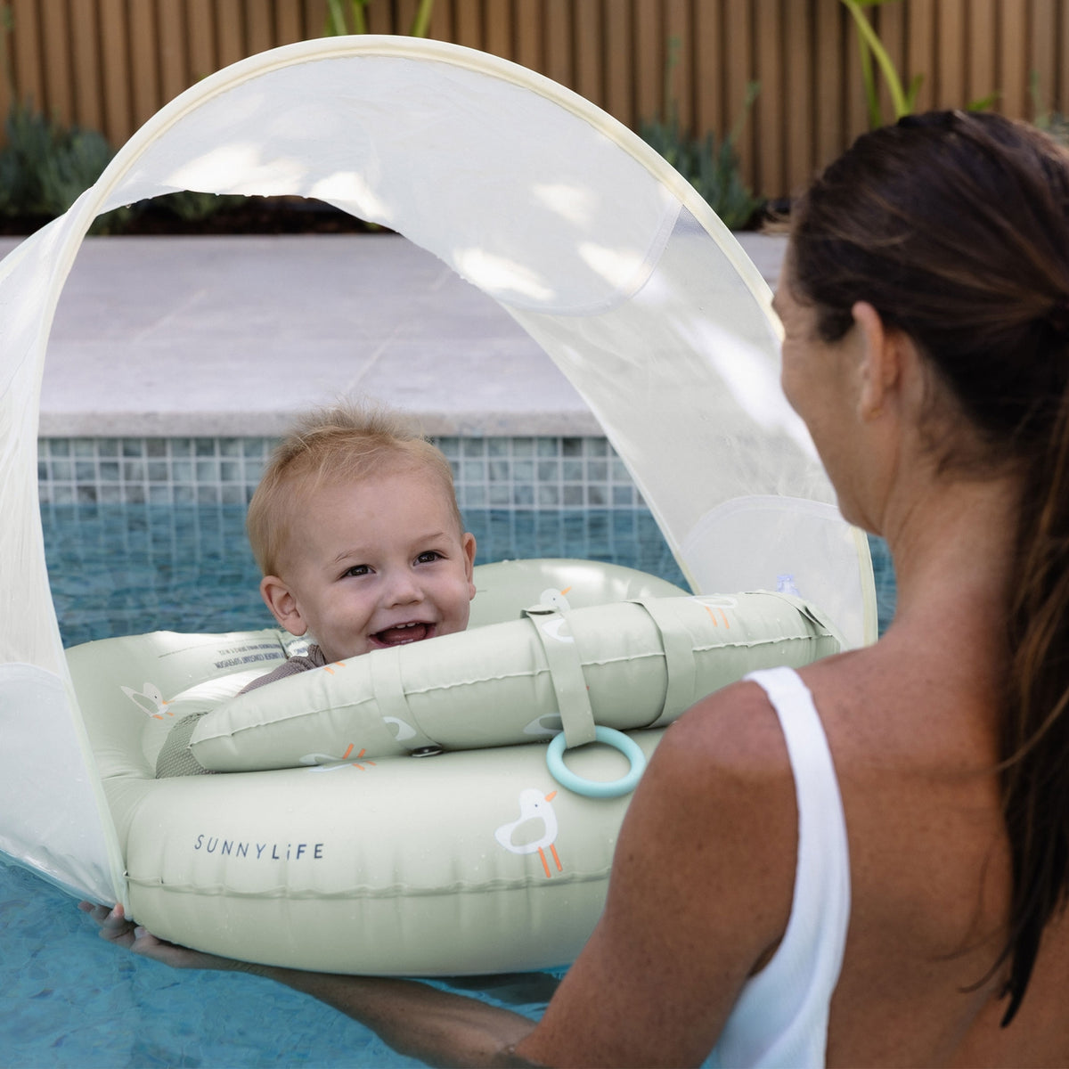 Child in a pool with a Sunnylife inflatable ring and canopy, watched by a woman.