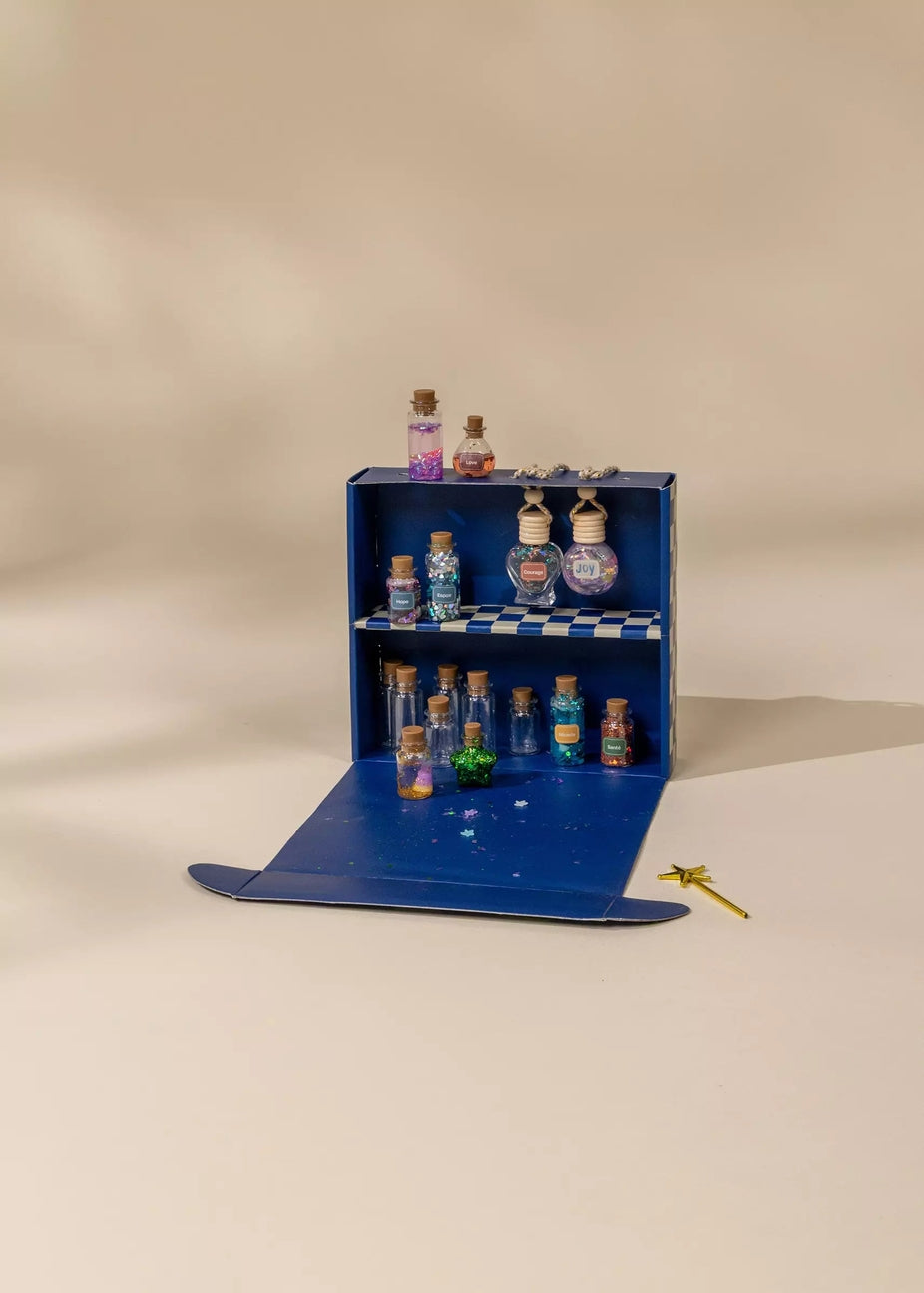 A miniature blue cardboard display shelf with tiny glass vials and bottles, set on a beige studio background with a small gold dragonfly prop nearby.