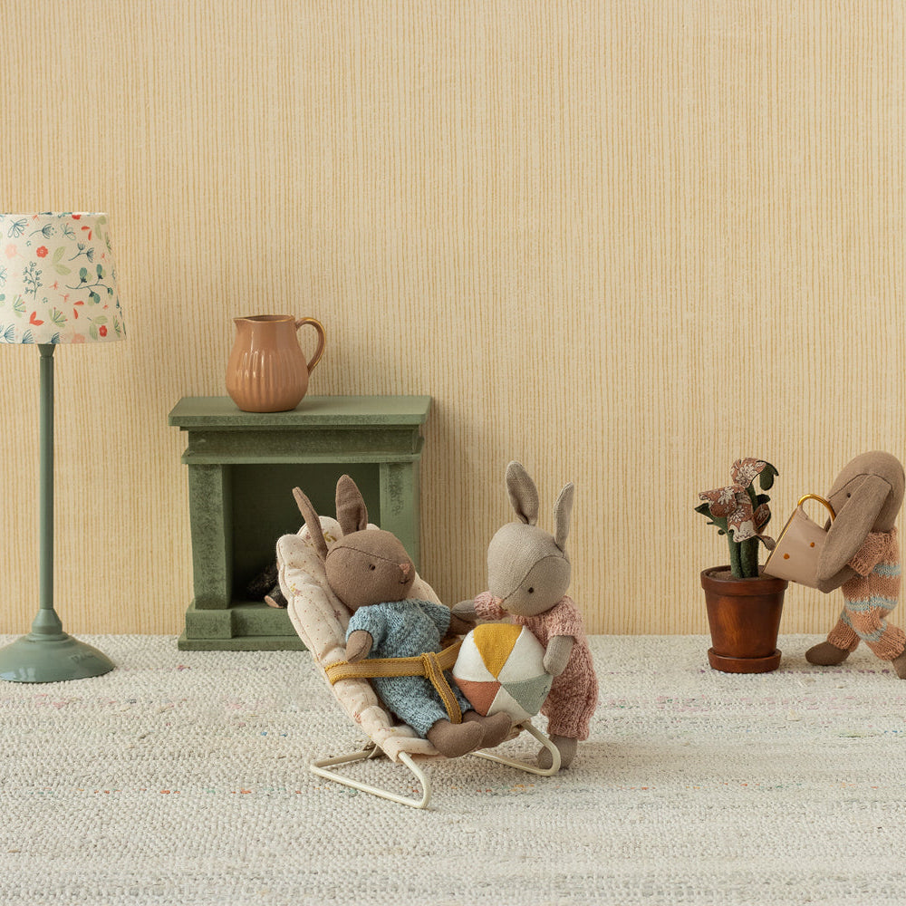 Two toy Maileg micro plush rabbits in a nursery scene, with one rabbit sitting in an off-white bouncy chair holding a small ball while another rabbit stands nearby.
