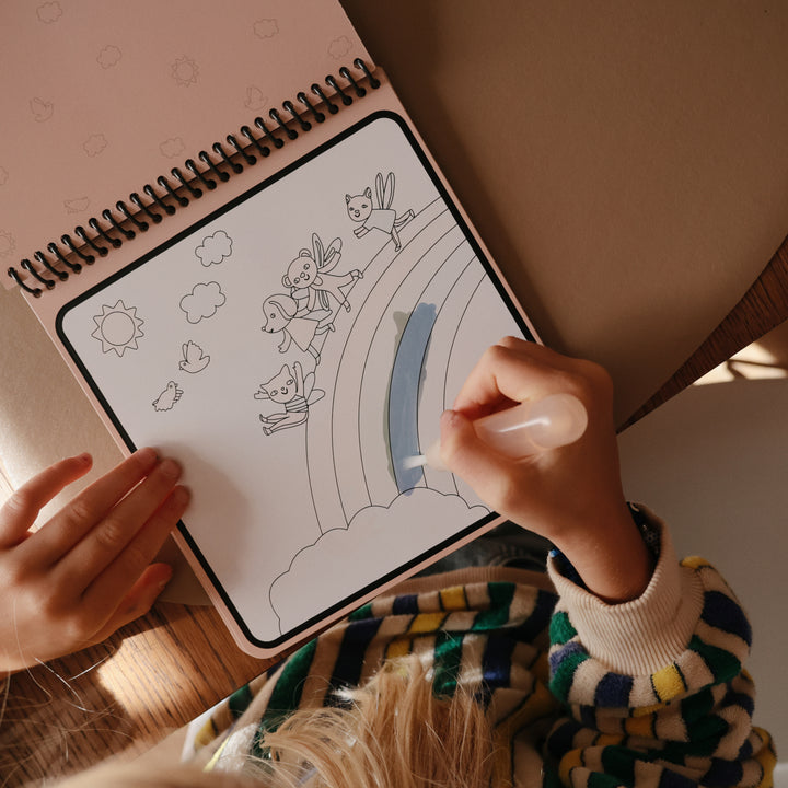 Person coloring a rainbow with a fairy in a spiral-bound coloring book.