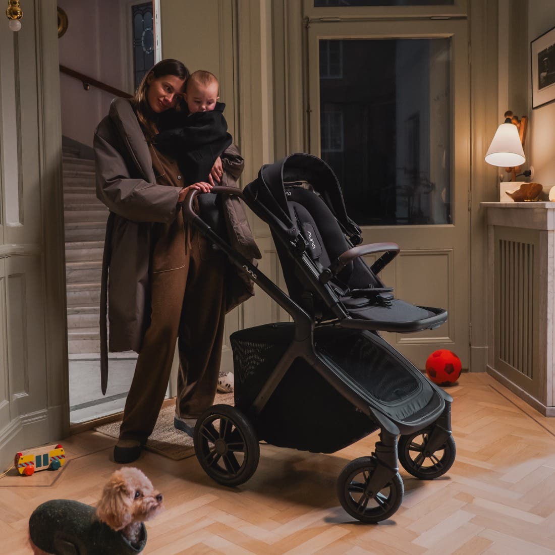 Woman holding a baby next to a black stroller in a home setting with a dog and toys on the floor.