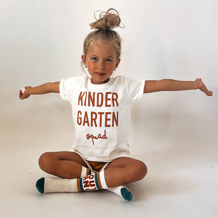 A young girl with her hair in a high bun sits cross-legged with arms outstretched, wearing a white t-shirt that reads 'KINDER GARTEN squad' in brown lettering, brown shorts, and white knee-high socks with brown and teal accents.