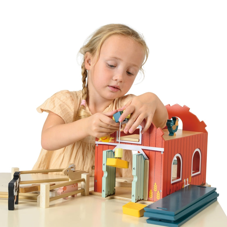 A young girl with blonde braids, wearing a short-sleeved yellow dress, plays with the Wooden Big Red Barn Playset by Mentari, holding a wooden toy horse near the open barn doors and fenced enclosure.