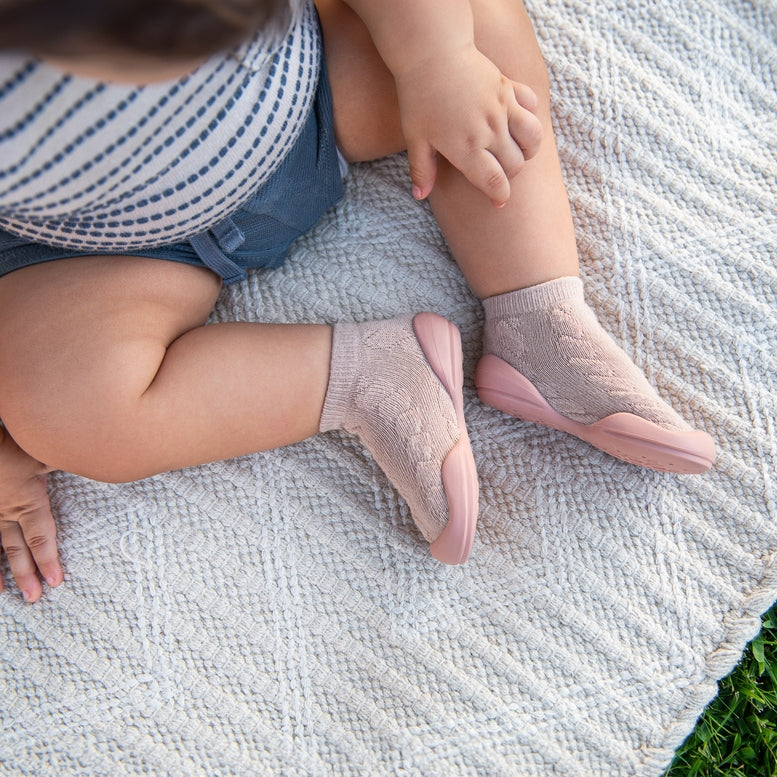 A close-up lifestyle shot of a baby sitting on a textured cream knit blanket outdoors, wearing light pink knit socks with solid pink toes.