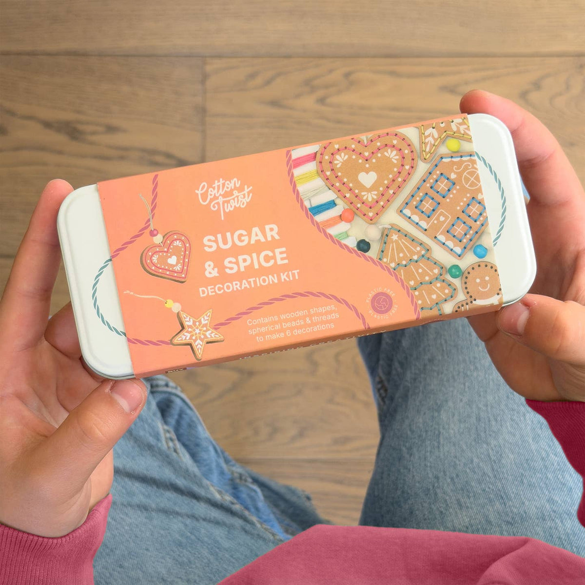 A person holding a Cotton Taster Sugar & Spice Decoration Kit tin, showing the packaging with gingerbread-themed illustrations against a wooden floor background.
