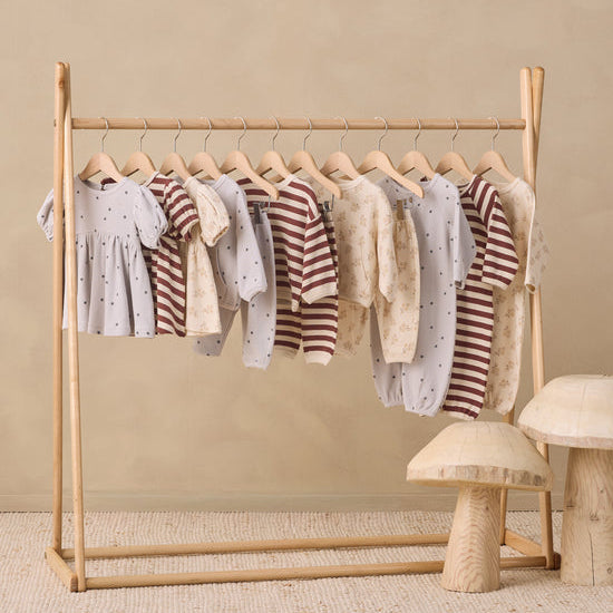 A wooden baby clothing rack holding multiple baby garments in soft neutral tones, set against a warm beige wall with a textured rug and small mushroom-shaped side tables.