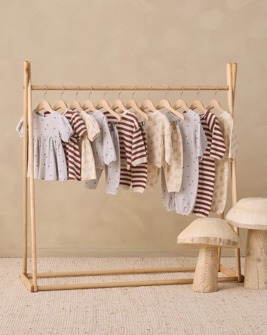 A wooden baby clothing rack holding multiple baby garments in soft neutral tones, set against a warm beige wall with a textured rug and small mushroom-shaped side tables.