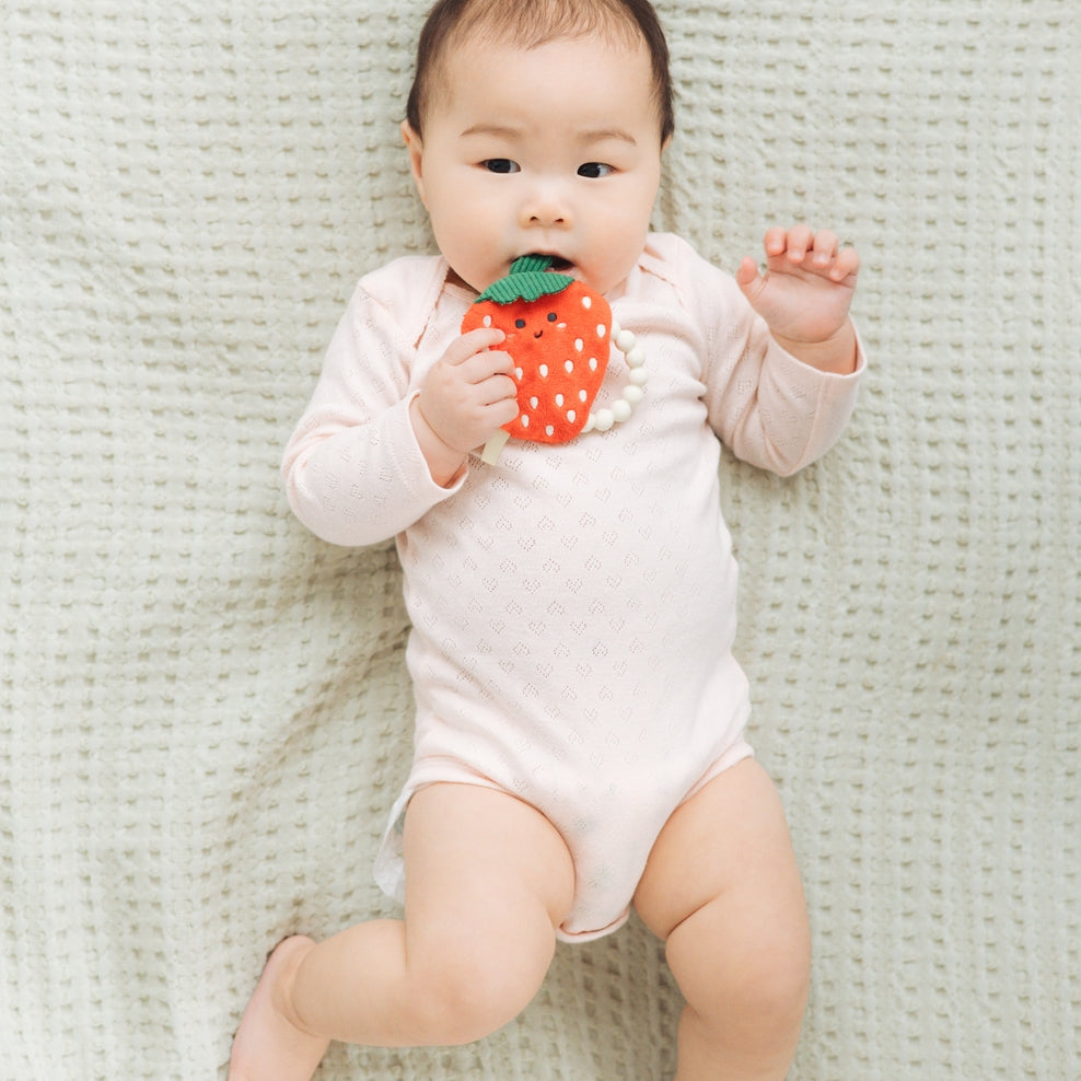 Baby in a white onesie holding a strawberry-shaped toy on a light green textured blanket