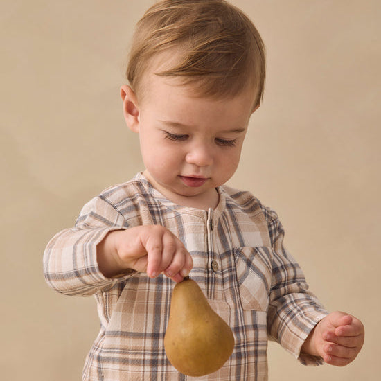 A toddler boy wearing a beige and brown plaid long-sleeve shirt holds a brown pear against a warm beige studio background.