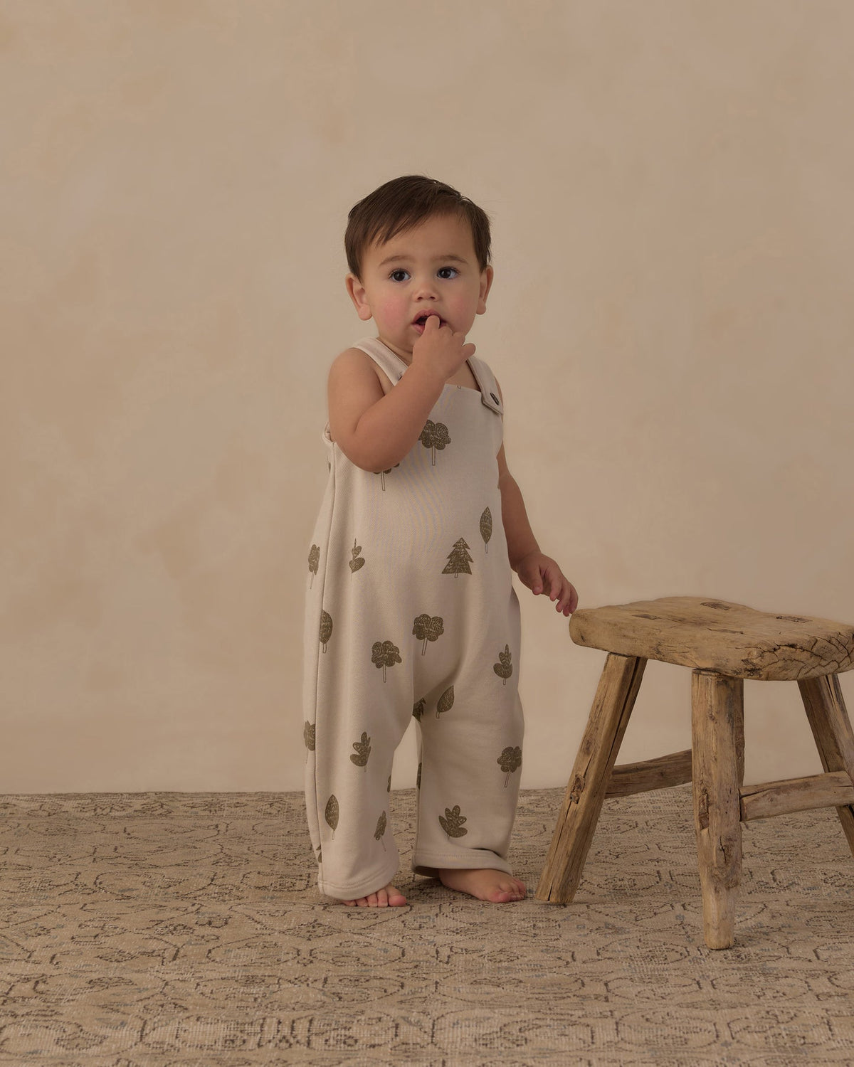 A baby standing on a textured rug next to a rustic wooden stool, wearing a cream-colored sleeveless romper with a green tree pattern, set against a warm beige studio backdrop.