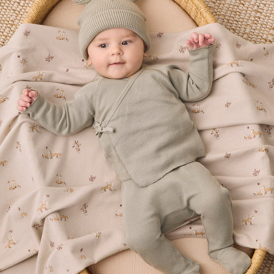 A baby wearing a sage green knit footed one-piece outfit with a matching beanie hat lies on a beige blanket printed with small deer, inside a round woven basket.