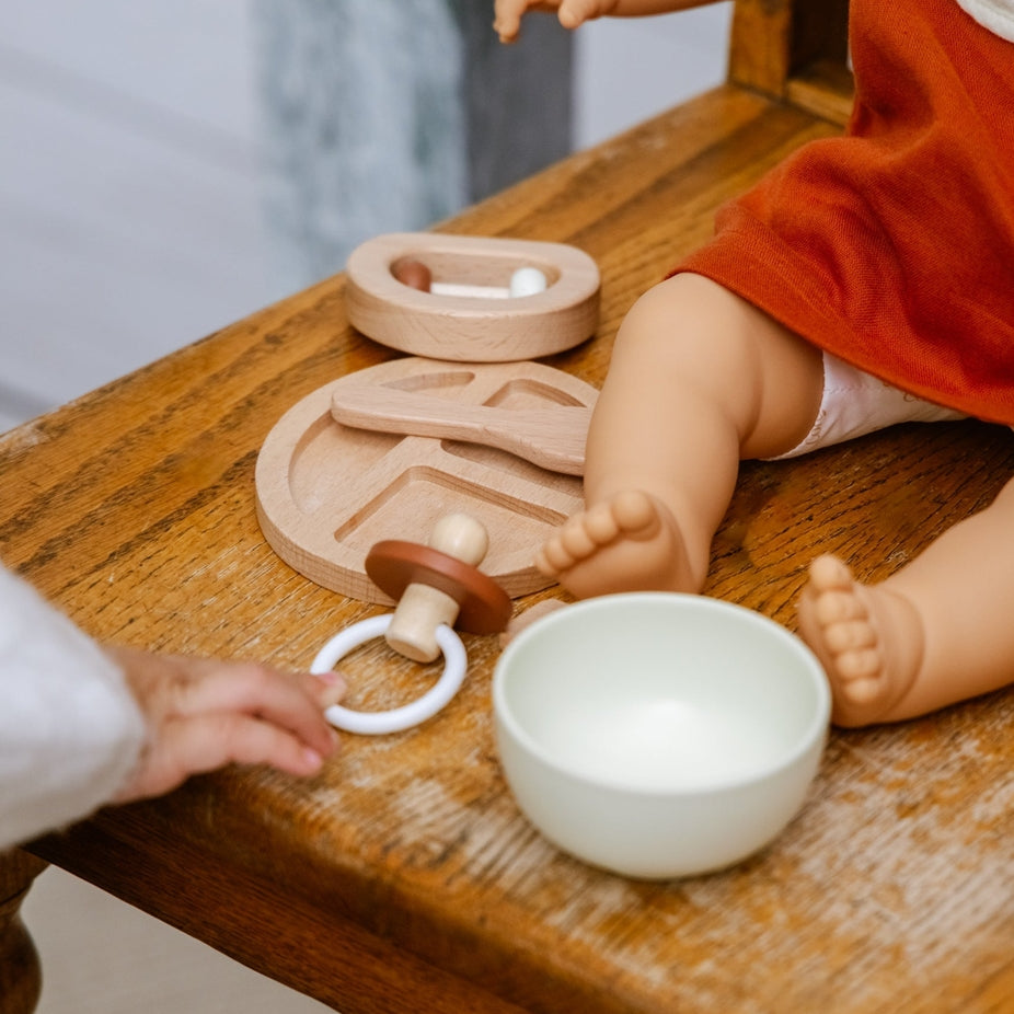 A close-up of a wooden baby sensory toy set on a rustic wooden table, featuring a wooden rattle, a wooden teething ring with a white ring, a small wooden spoon, and a white ceramic bowl. A doll in an orange dress and a baby's hand are interacting with the toys.