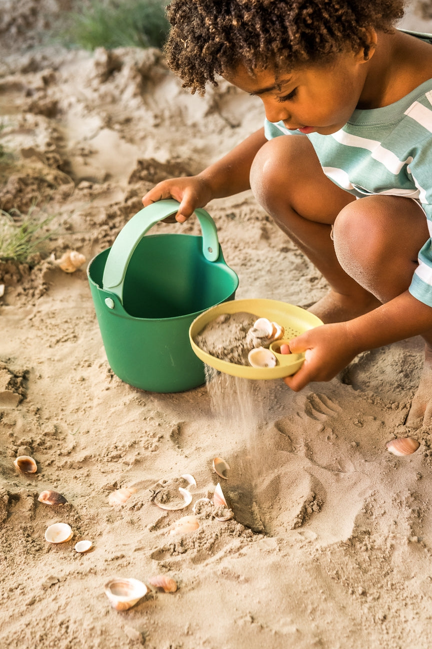 Child playing with sand at the beach using a green bucket and yellow bowl.