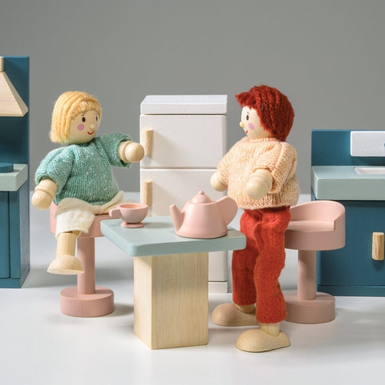 Wooden kitchen furniture set by Mentari against a white background, featuring two wooden dolls seated at a light wood-colored table with a teapot and two cups, surrounded by a blue stove with an oven and hood, a white refrigerator with wooden handles, and a blue sink unit with a cupboard.