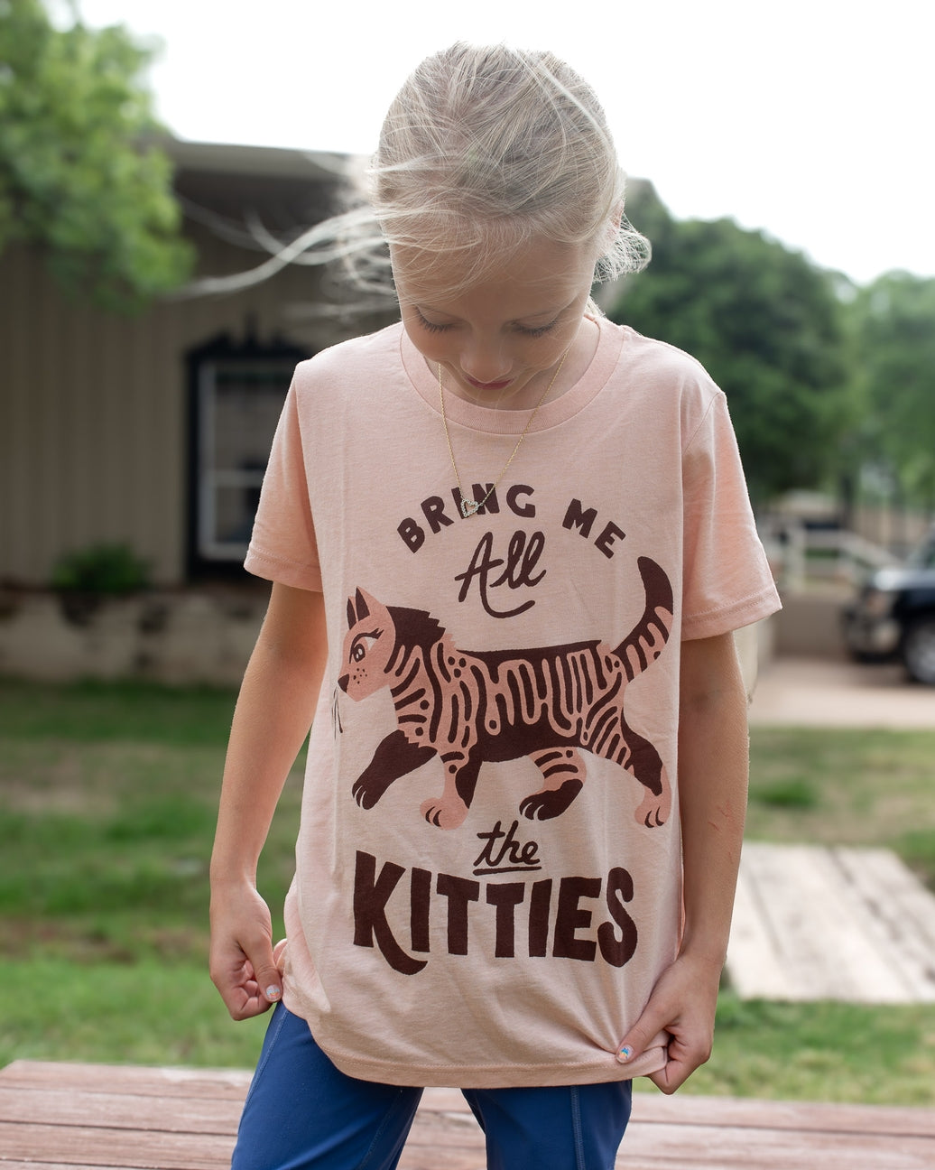 A young girl wearing a light peach-pink graphic t-shirt with a brown cartoon kitten and the text 'BRING ME All the KITTIES'. She is standing outdoors on a wooden deck with a blurred house and greenery in the background.