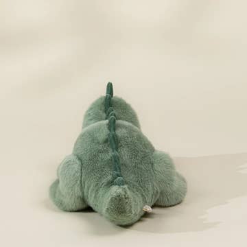 A plush dinosaur toy in a dusty sage green color, photographed from behind against a neutral, light beige background.