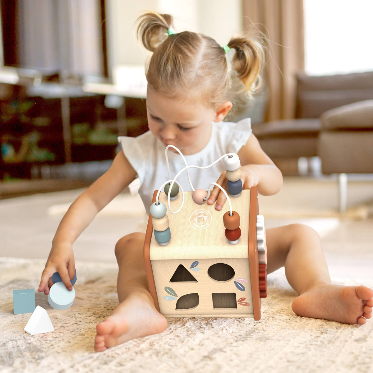 A toddler girl with pigtails sits on a light rug playing with a wooden shape-sorting activity box. The box has colorful pegs, beads, and geometric panels, and the child is surrounded by matching wooden blocks in a bright, softly blurred living room.