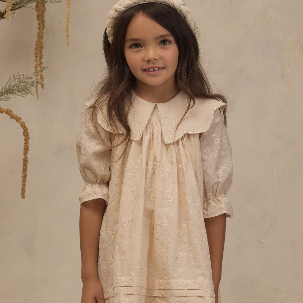A young girl with long brown hair wearing a cream-colored lace dress with a large Peter Pan collar, puff sleeves, and a matching braided headband, standing against a textured beige wall.