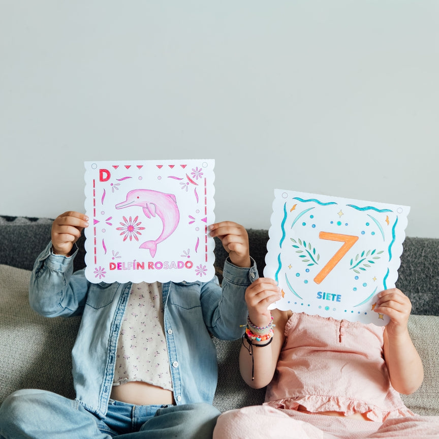 Two children sitting on a gray couch holding up colorful educational cards. The child on the left wears a denim jacket and blue jeans, holding a pink card with a dolphin illustration.