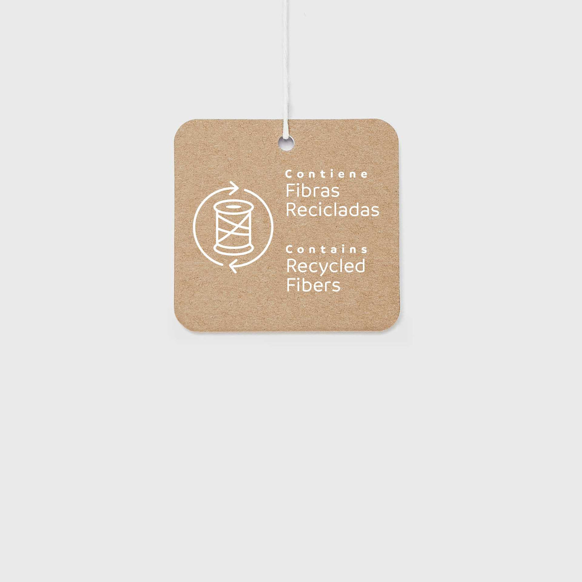 A square kraft paper tag with rounded corners, featuring white text that reads 'Contiene Fibras Recicladas / Contains Recycled Fibers' alongside a recycling symbol, suspended by a thin white string against a light gray background.