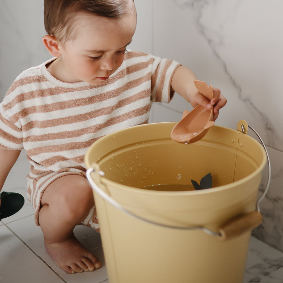 A young child in a striped shirt sitting on a tiled floor while playing with a yellow bucket of water and a toy whale.