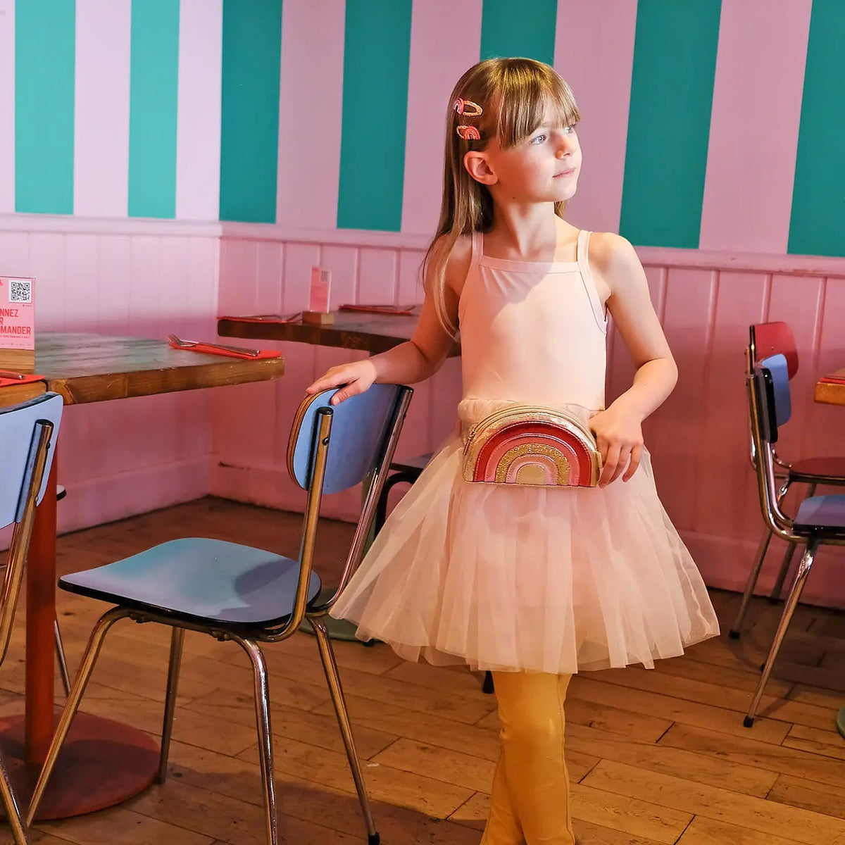 Young girl in a pink dress holding a colorful bag in a room with striped walls.