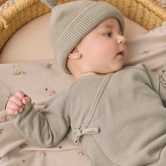 A baby lying on a patterned blanket inside a woven bassinet, wearing a textured sage green knit hat with a small pom-pom and a matching sage green long-sleeve romper.